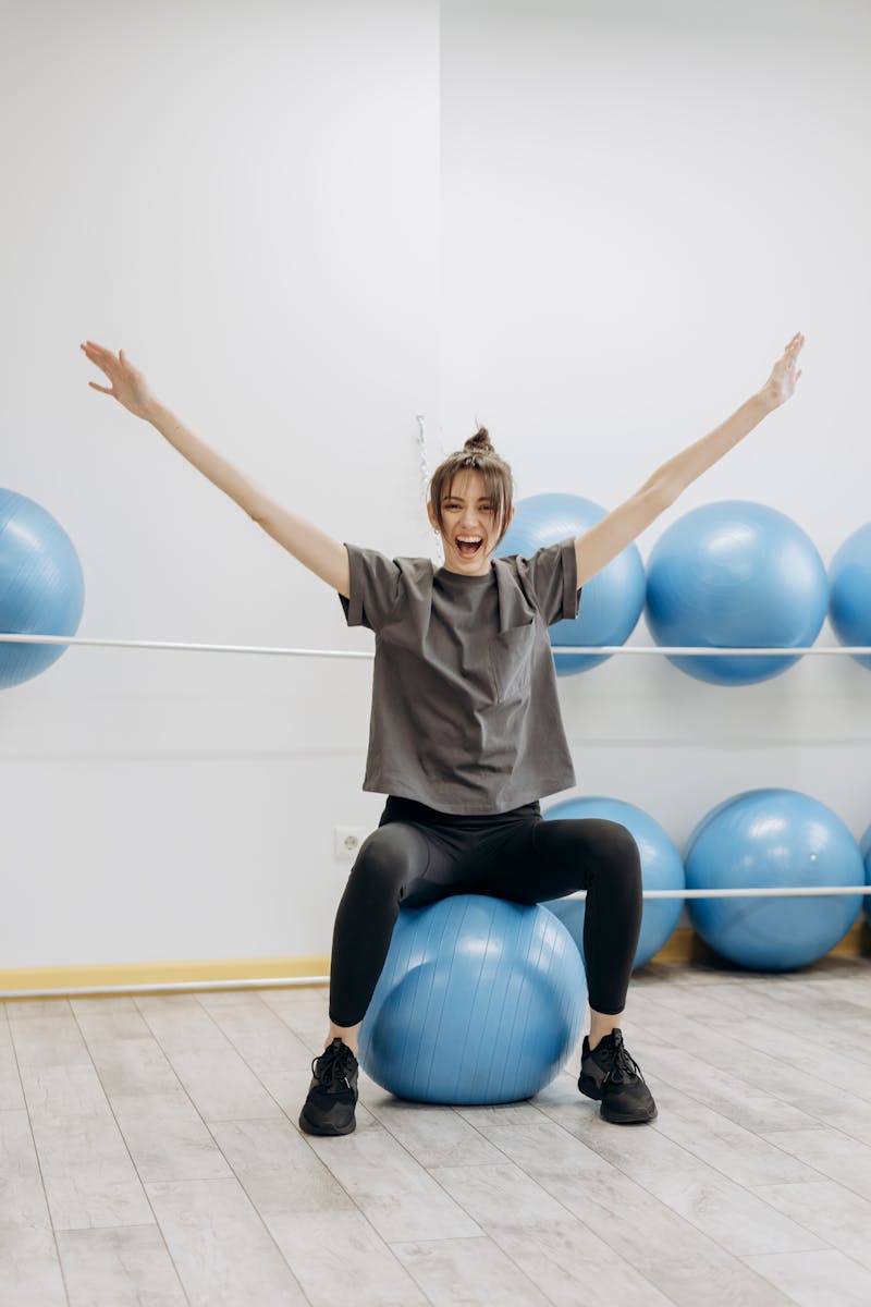 Photo by Mikhail Nilov A cheerful woman in active wear exercises with a yoga ball indoors, promoting wellness.